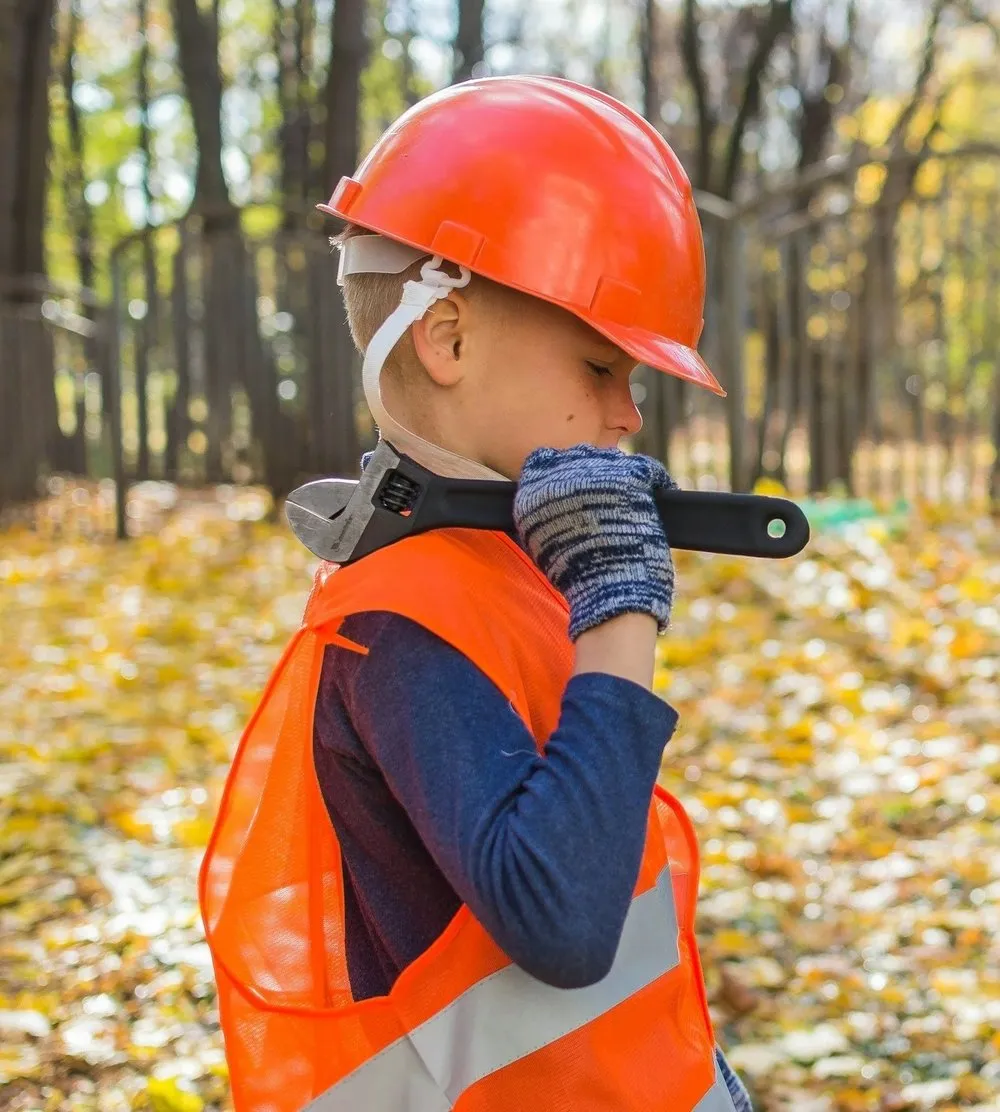 A child in a hard hat and orange vest carrying a wrench outdoors